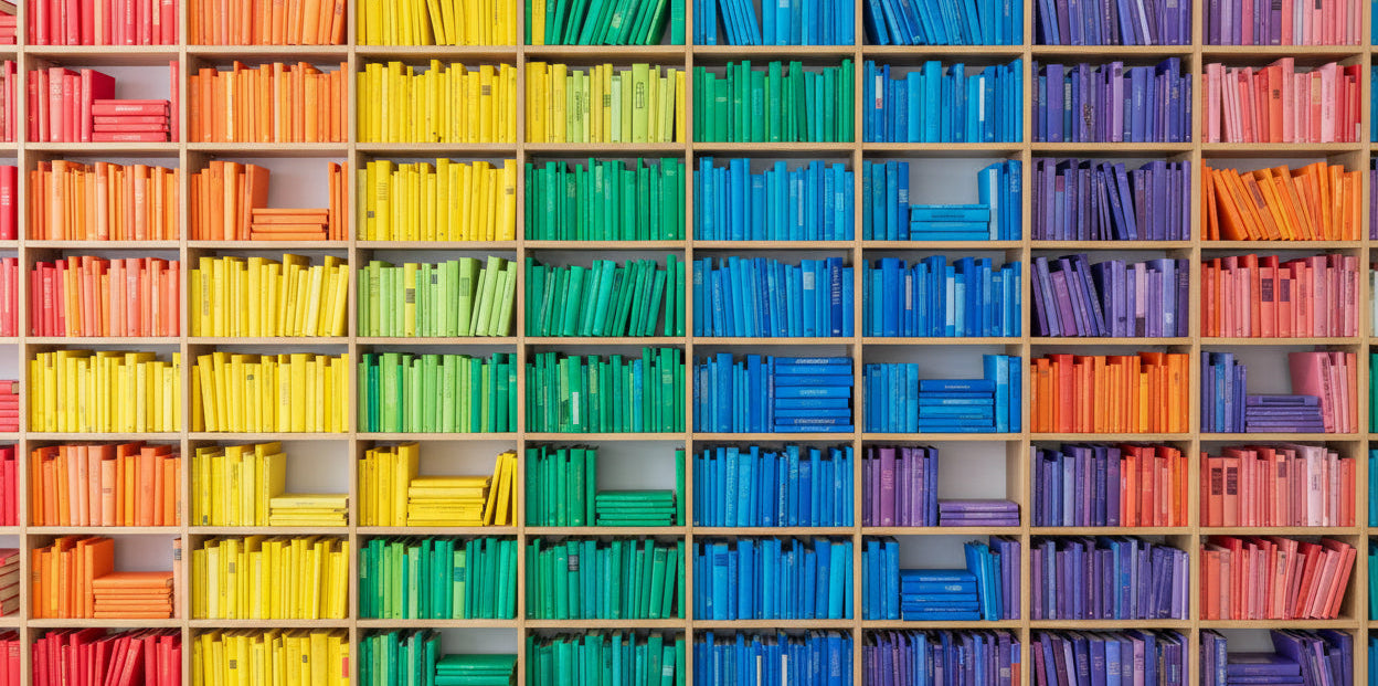 Wooden wall shelves filled with brightly colored books arranged in vertical and horizontal color blocks of blue, purple, yellow, pink, orange, green, and teal.